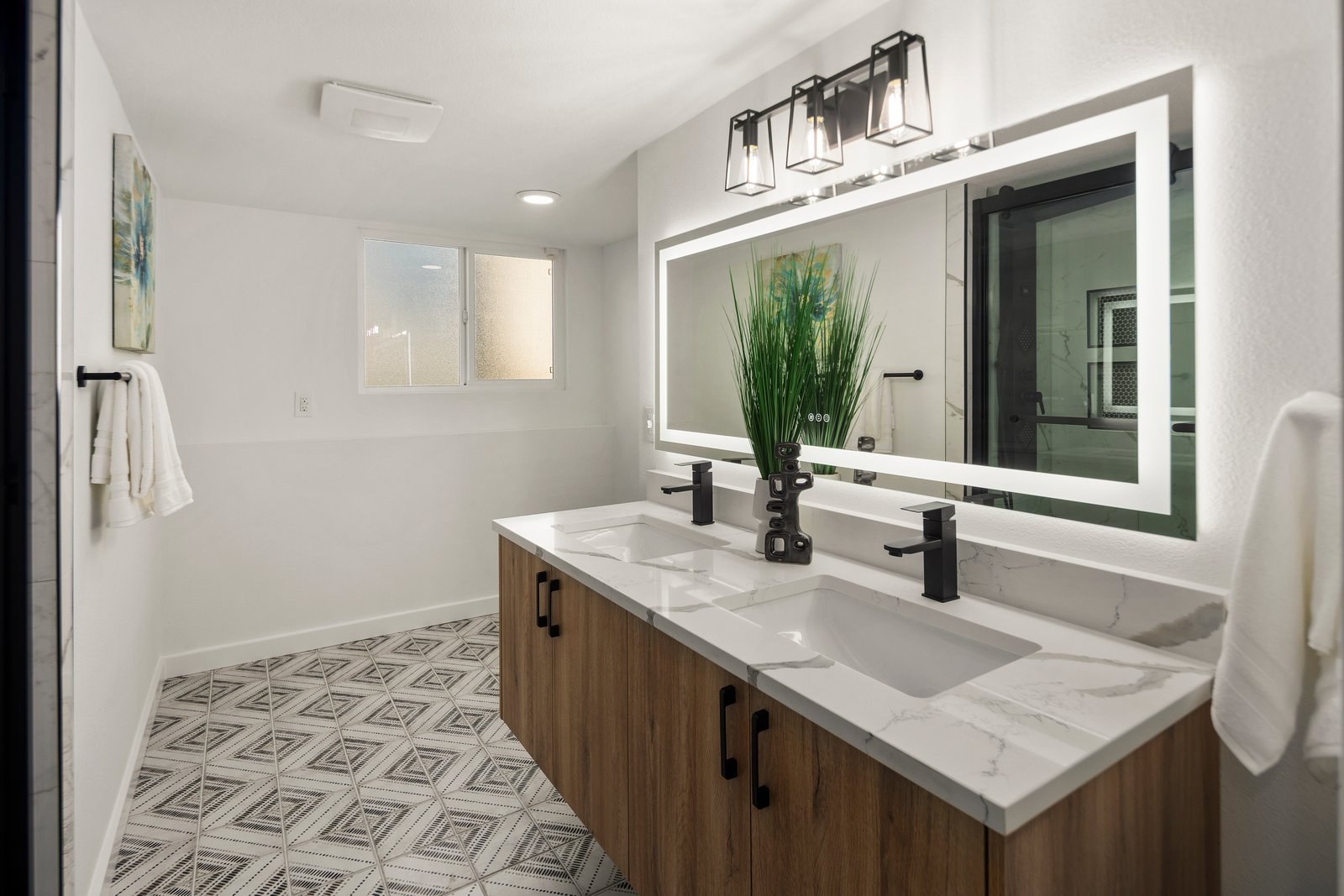 Renovated bathroom with double vanity, lit mirror and patterned tile floor